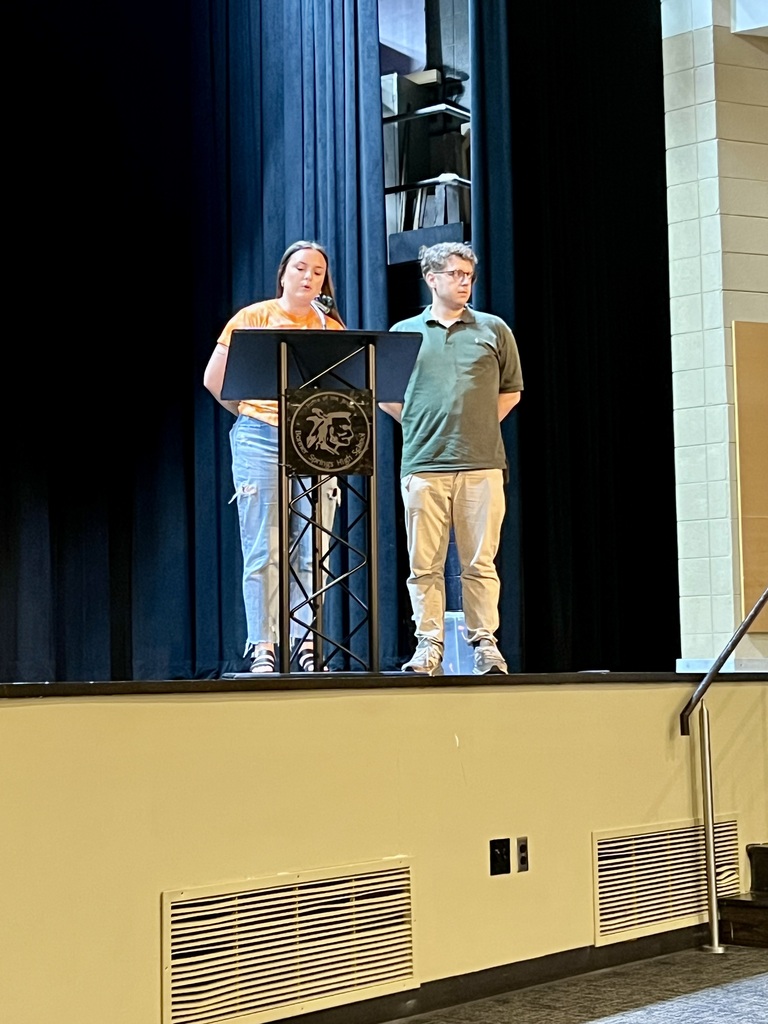 Two individuals stand on a stage with a podium, one wearing a green shirt and the other an orange top.
