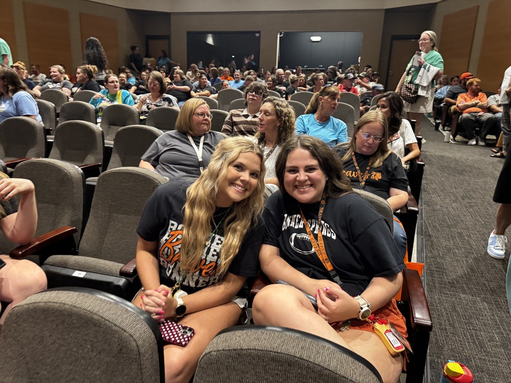 Two women sit in a large room with many people seated in rows of chairs.