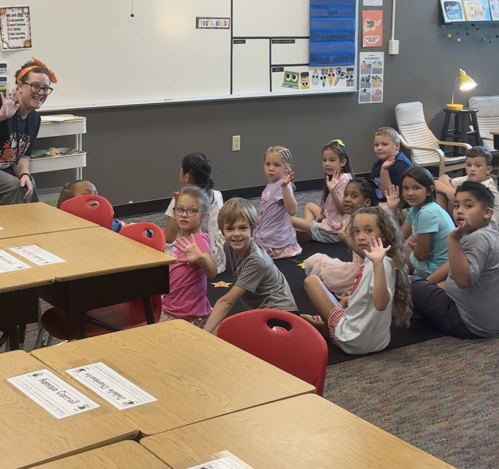 A classroom with children seated in chairs. Some are standing and raising their hands. A person stands in front, addressing them.