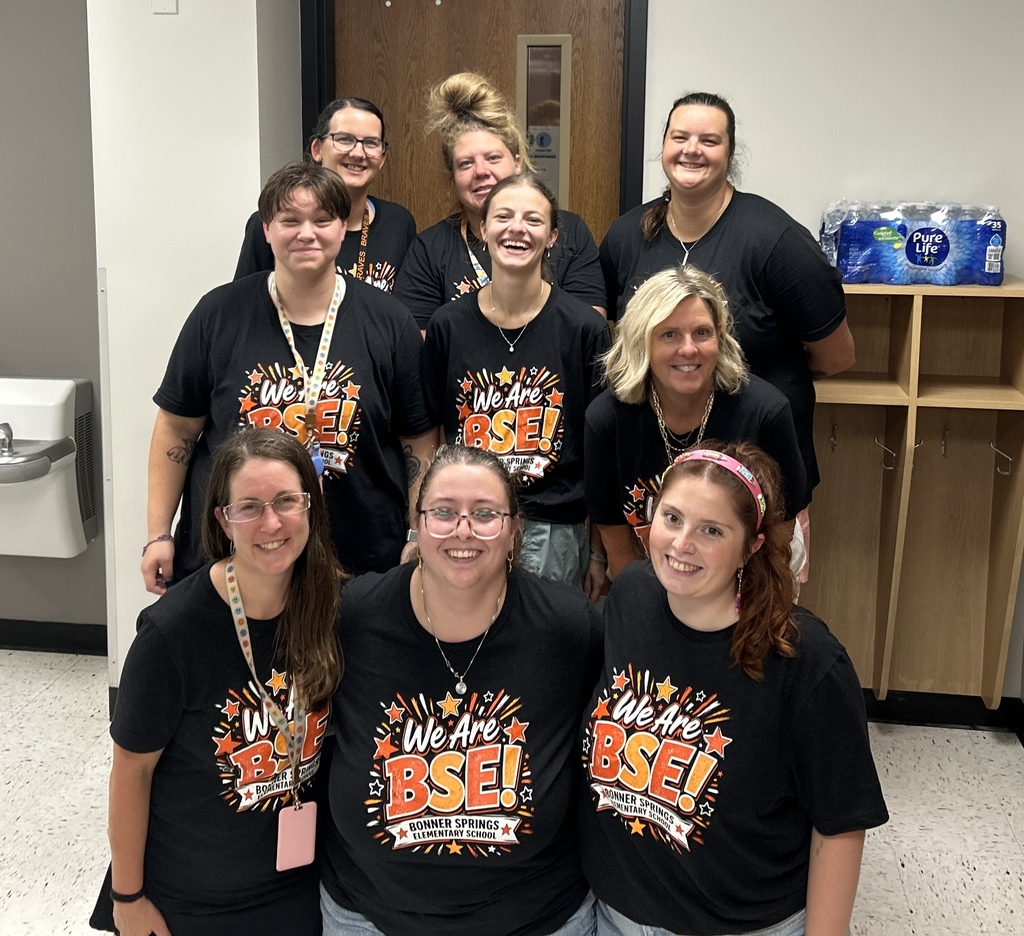 A group of women wearing matching black t-shirts stand in a hallway. Some wear glasses, and some wear necklaces.