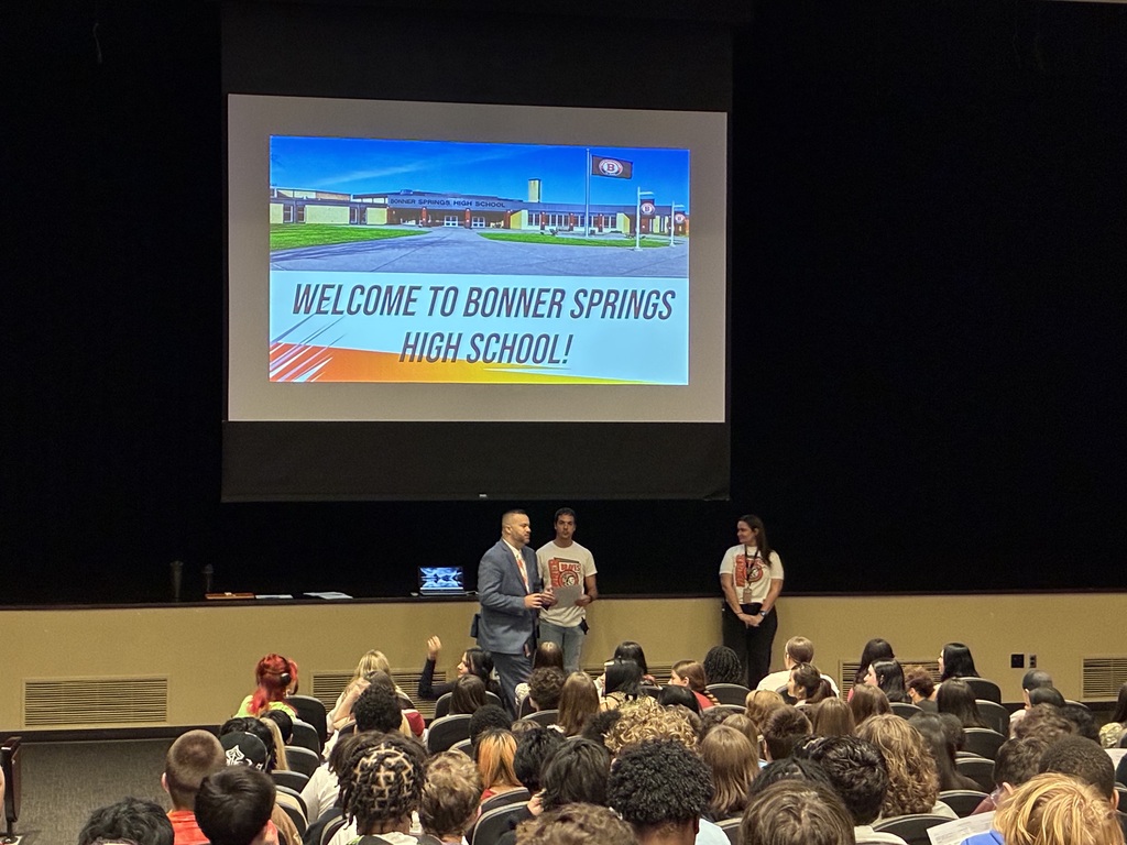 Three people stand on stage in front of an audience, with a large screen displaying the message "Welcome to Bonner Springs High School."