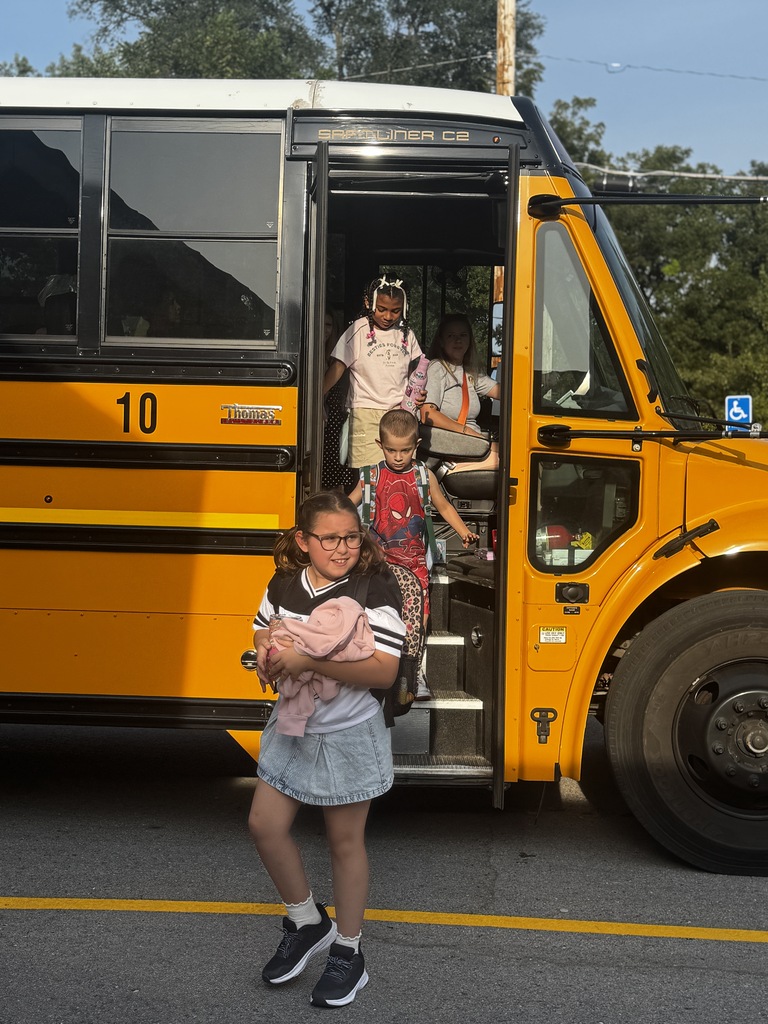 A school bus is open and children are inside. A girl in glasses stands in front of the bus.