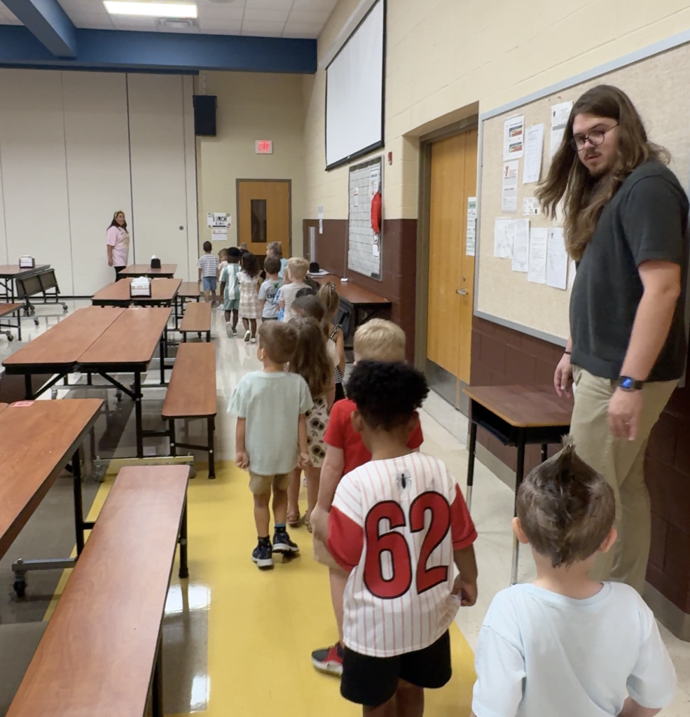 A man with long hair and a watch walks beside a line of children. The children walk past tables.