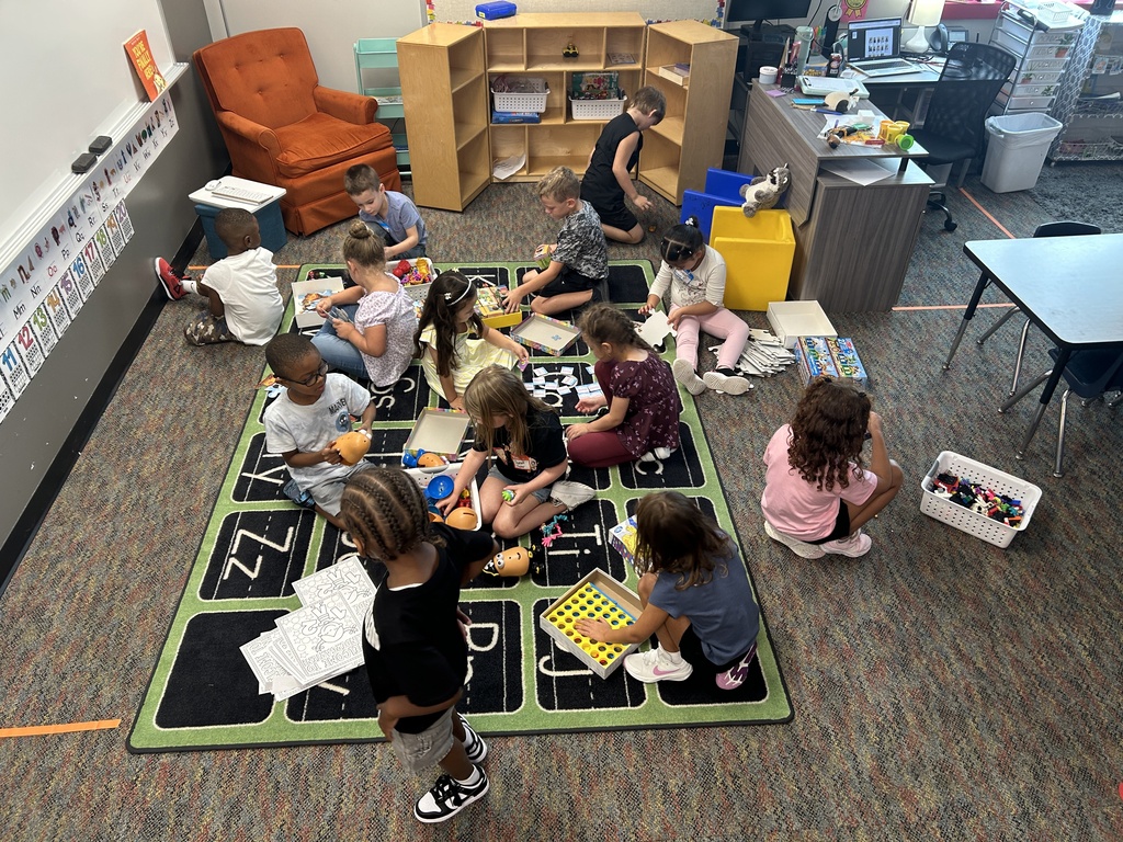 Children seated around a carpet with letters and numbers, engaging in an activity. Some children are holding objects. Background includes a classroom with shelves and chairs.