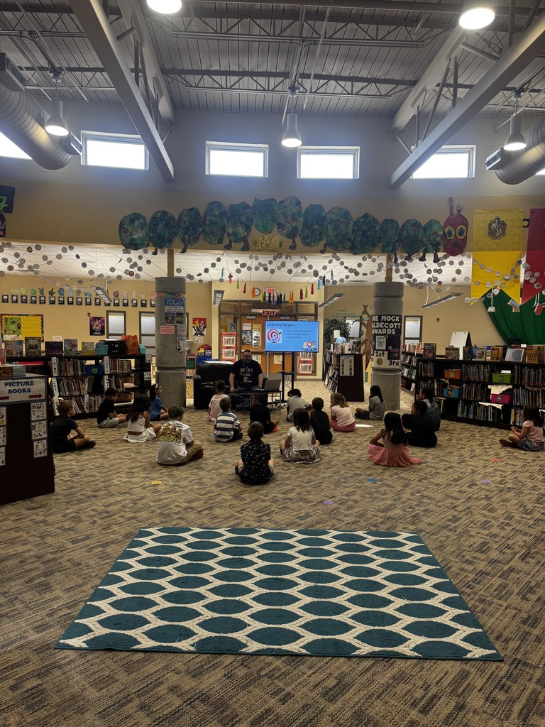A classroom with students seated on a carpeted floor. A person stands in front of a screen. Shelves filled with books and green foliage on the ceiling.