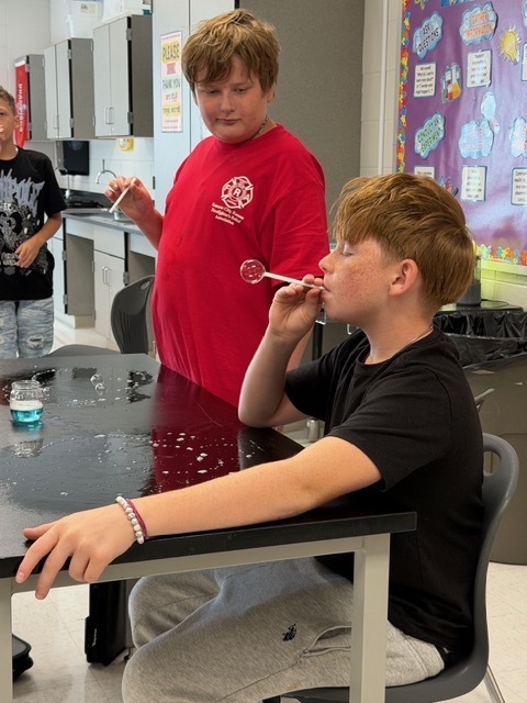 Two boys, one in red and one in black, blow bubbles with wands at a table. Behind them is a classroom with a bulletin board.