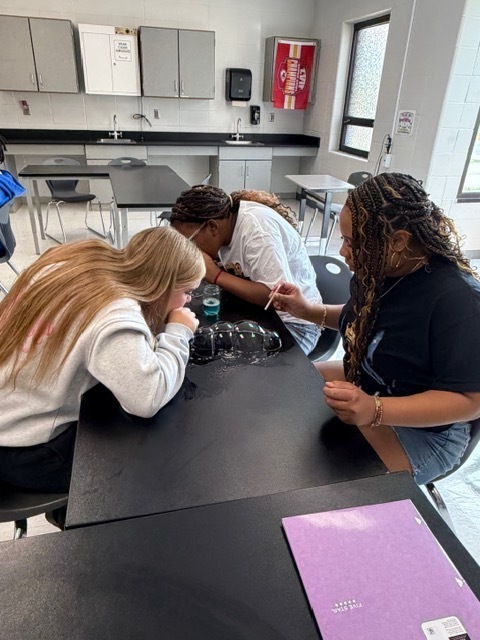 Three women in a classroom setting, two working on a lab table, one observing and using a pen.