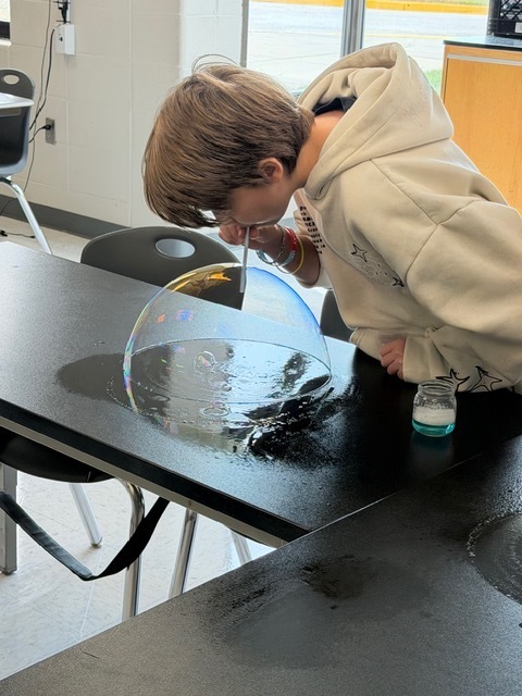 A child is blowing bubbles at a table, standing near a bowl of water. A sink and a chair are in the background.