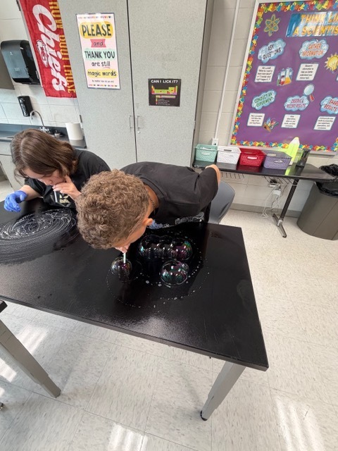Two individuals in blue gloves work together at a table, one pouring a liquid into a black container.