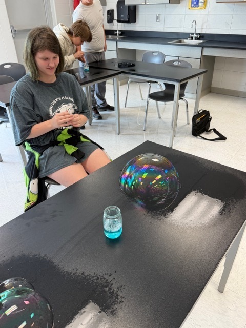 Woman seated at a table with a large, colorful bubble in the foreground and a small container with blue liquid on the table.