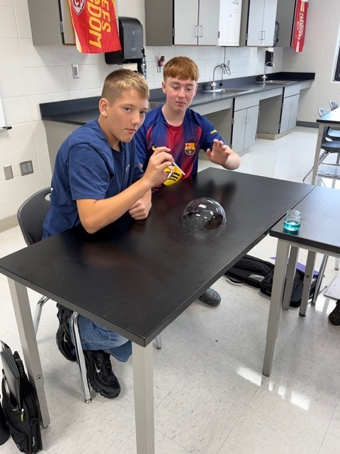 Two boys in a classroom, seated at a table with a ball on it, interacting with objects.