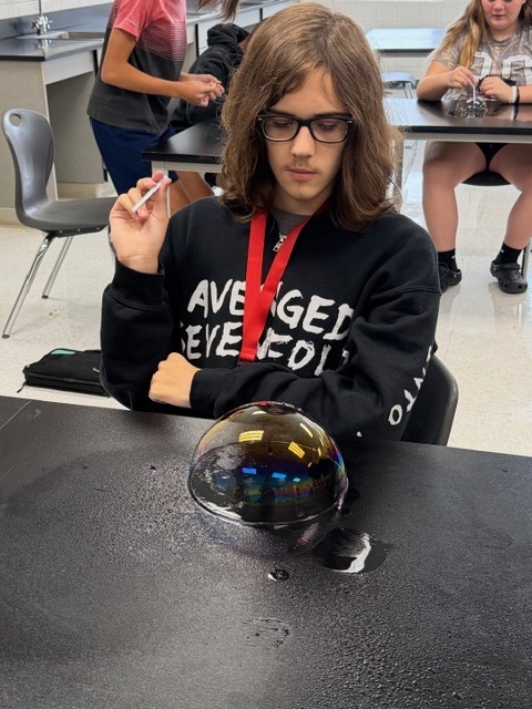 A student wearing glasses and a black sweatshirt uses a cigarette lighter to manipulate a transparent sphere on a table.