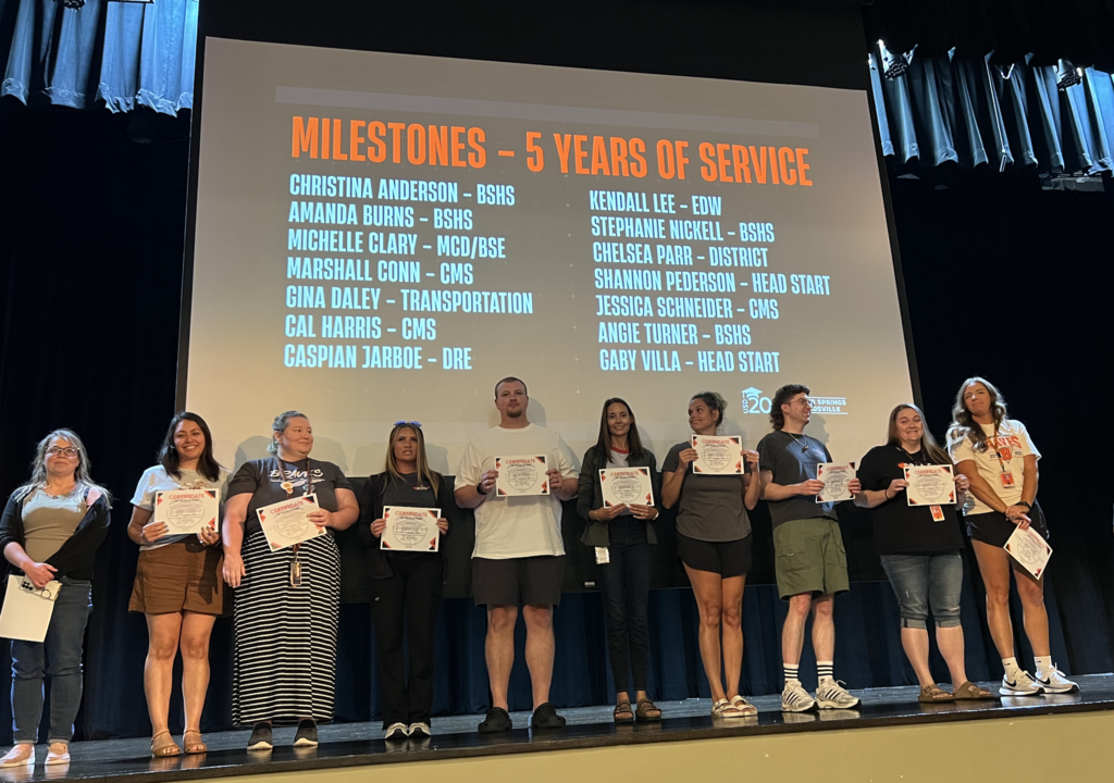 People holding certificates on stage in front of a screen displaying a list of names and years of service.
