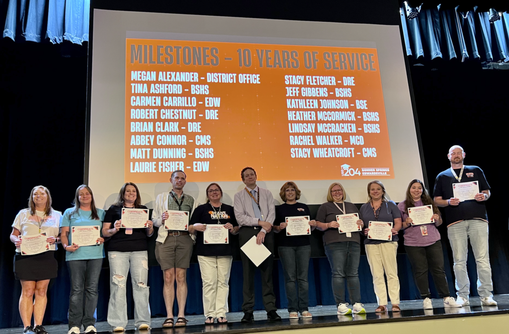 Group of adults on stage holding certificates. A screen behind them displays "Milestones - 10 years of service" with names.