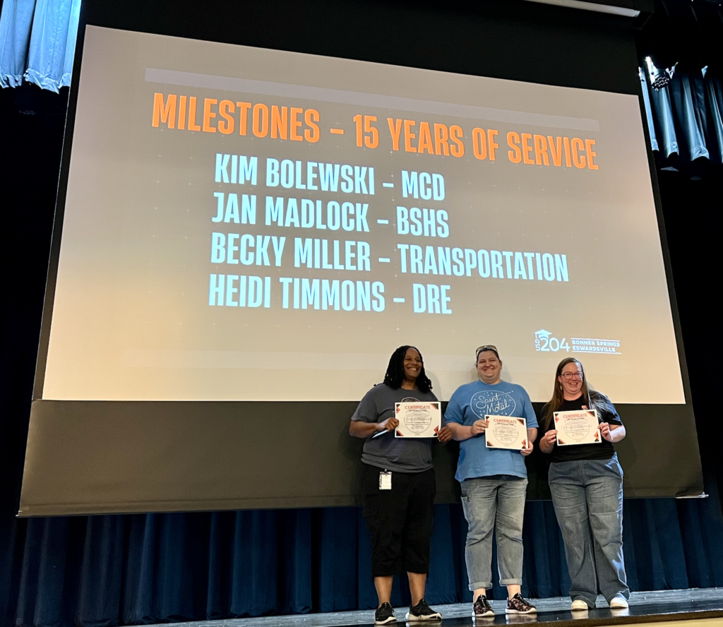Three individuals stand on a stage, holding certificates, with a screen behind them displaying milestones for 15 years of service.