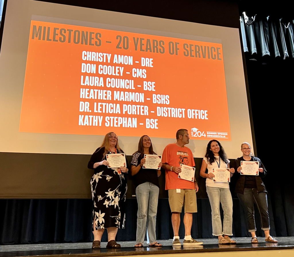 Five people stand on stage holding certificates. Behind them, a screen displays milestones and years of service.