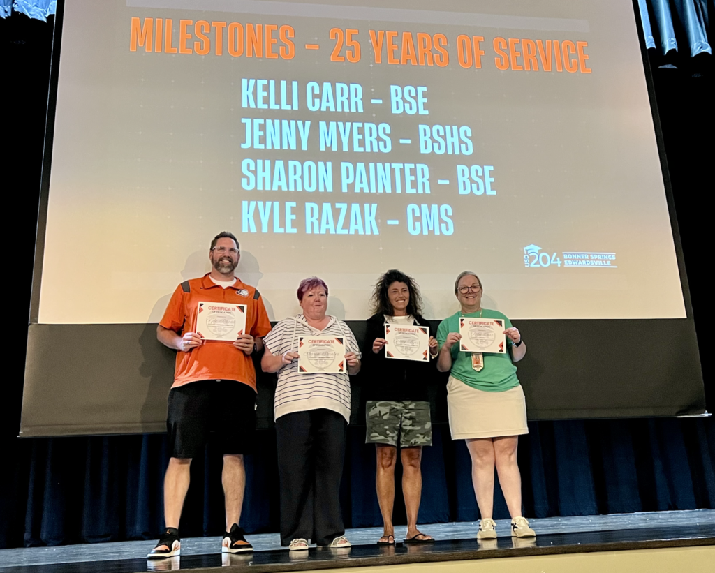 Five people stand on a stage holding certificates. A screen behind them displays "Milestones - 25 Years of Service" with names.