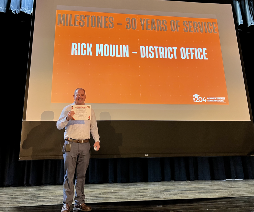 Person holding a certificate stands on stage in front of a screen displaying "Milestones - 30 Years of Service" text.