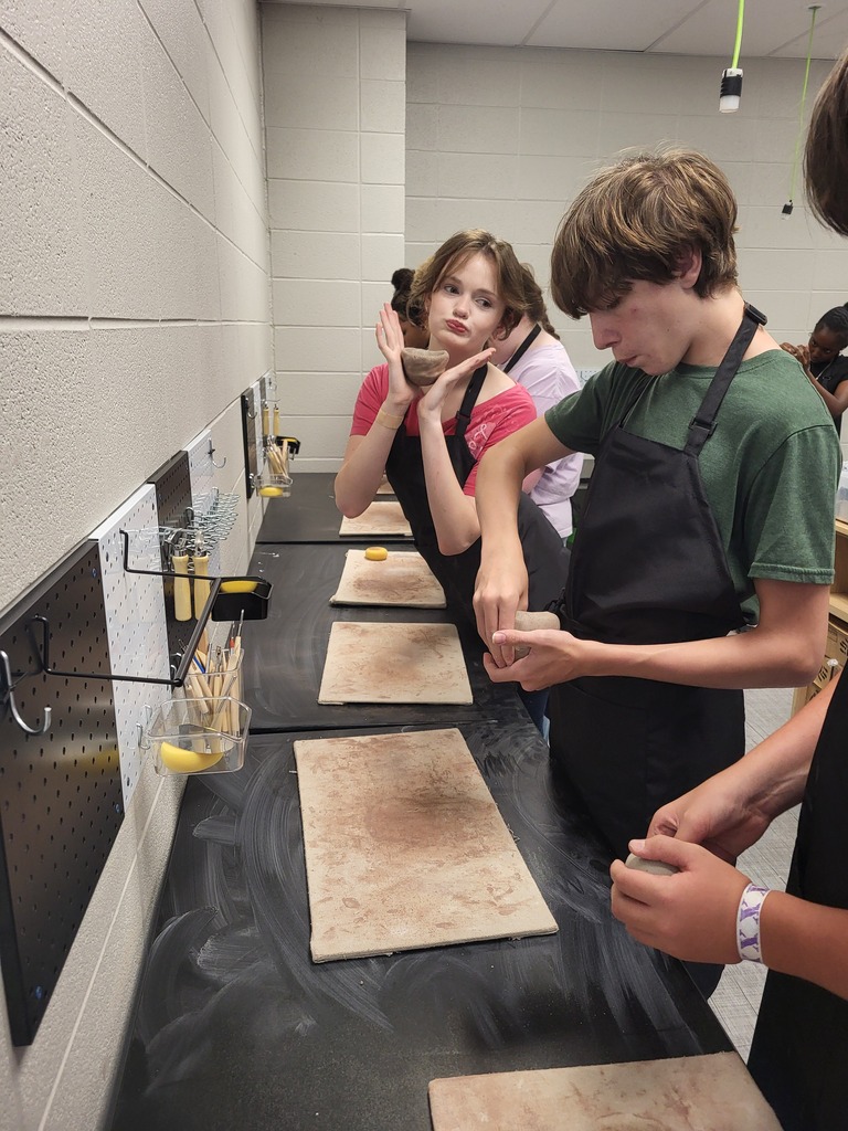 Several students in aprons work at a table, one holding a bowl and another a small object.
