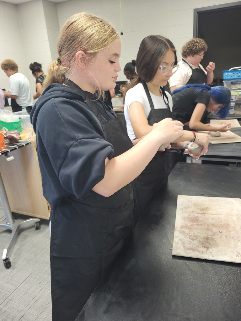 A woman in a black apron stands at a table in a classroom, working with materials.