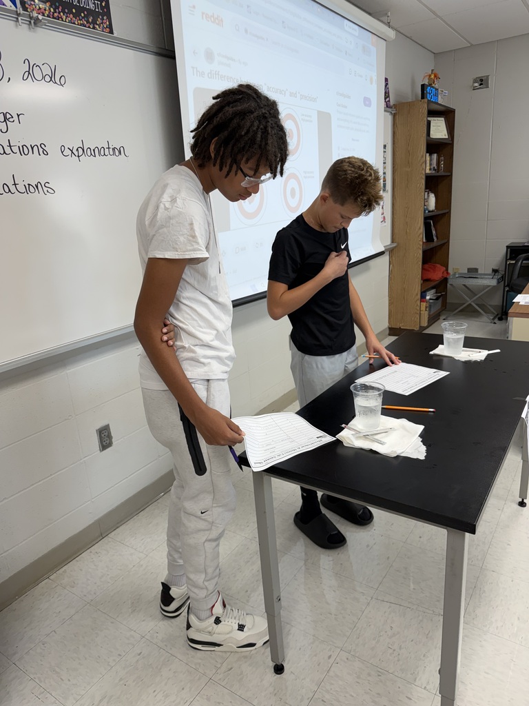 Two boys stand before a whiteboard in a classroom, one holding papers and a microphone.