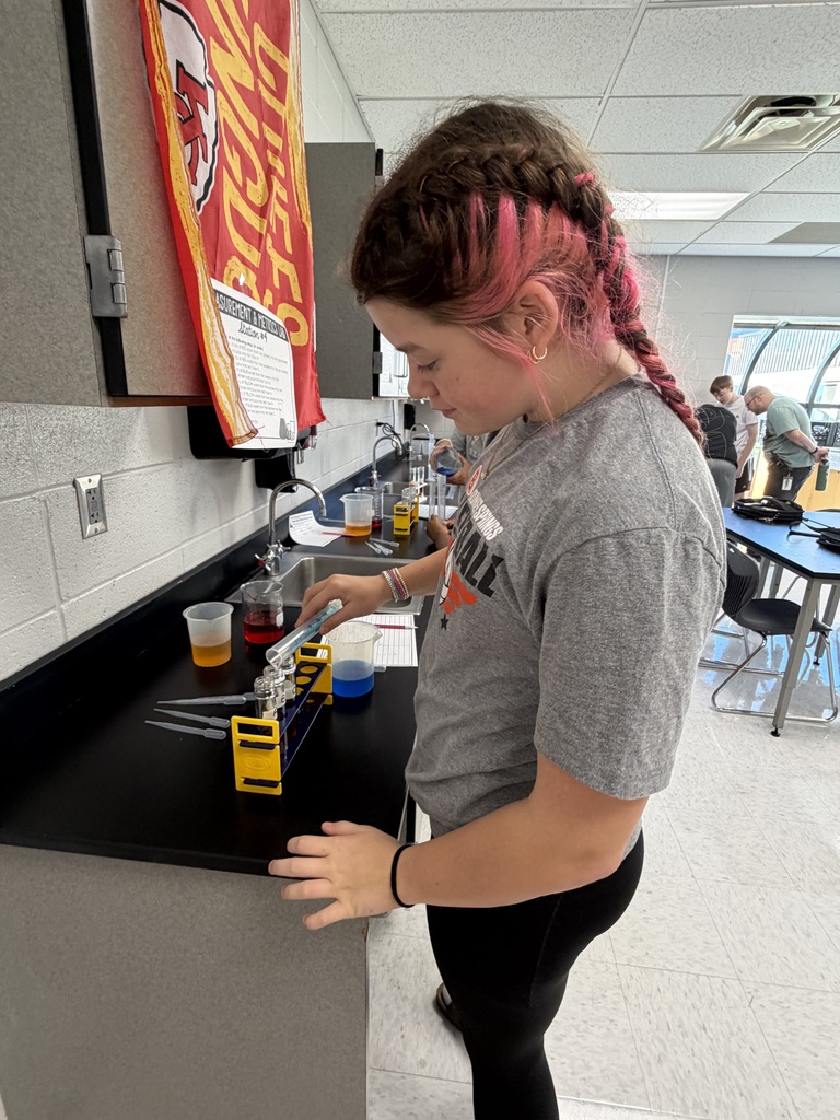 A woman in a gray shirt and pink hair braids prepares a chemistry experiment in a classroom.