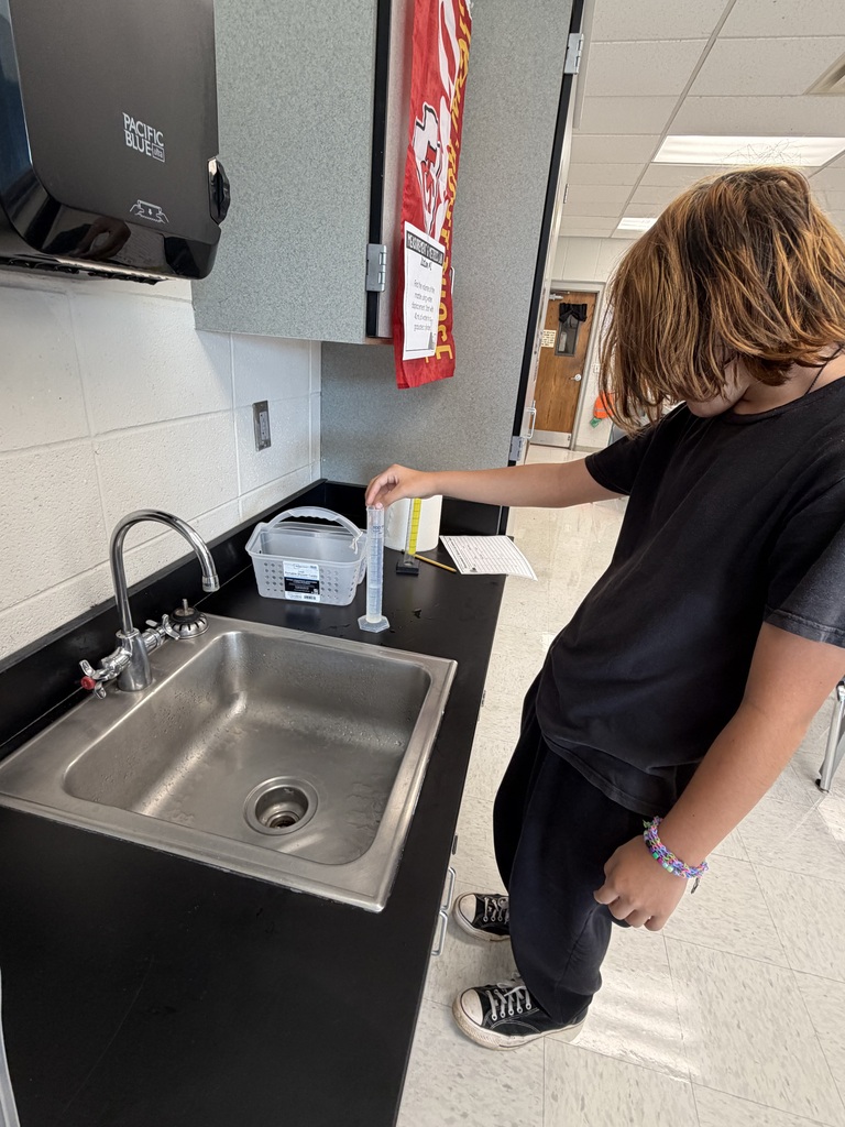 A person is standing at a sink, pouring water from a bottle into a clear measuring cup.