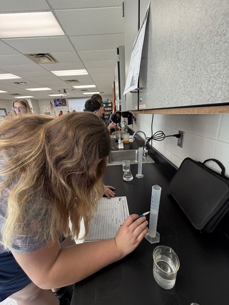 A student at a desk, using a pipette to fill a graduated cylinder with liquid.