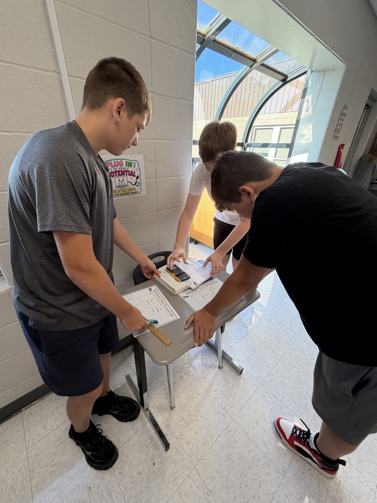 Three individuals in a hallway. One examines papers on a table while the others look on.
