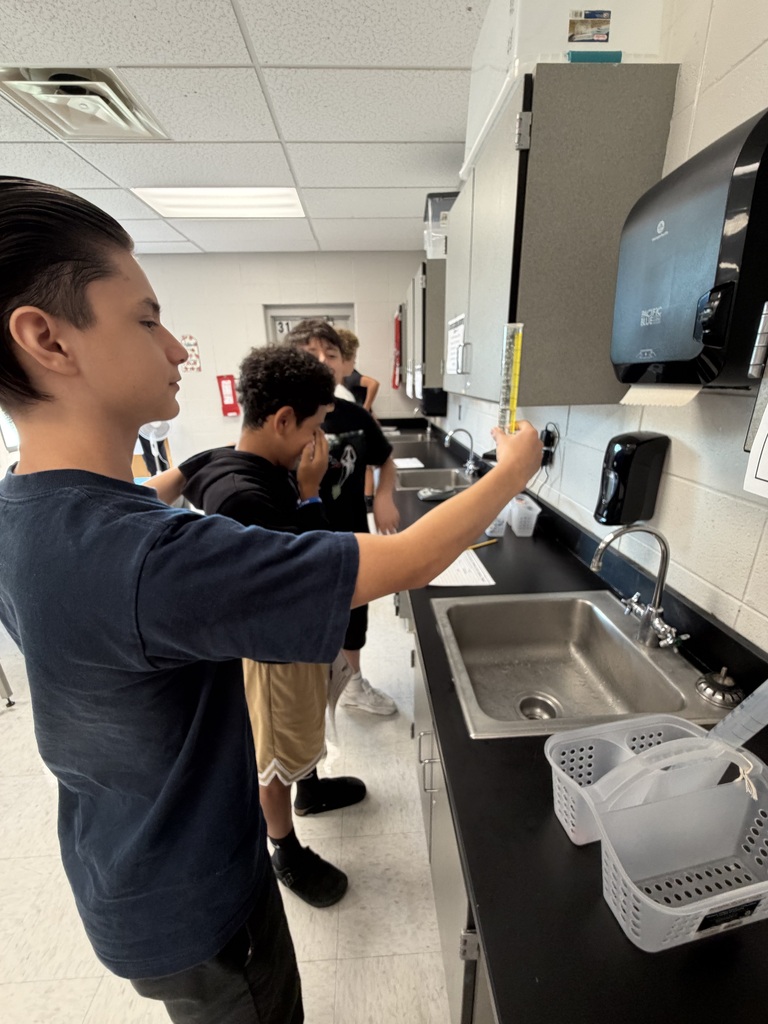 A person stands in a kitchen, pointing at a paper towel dispenser while others are behind him.