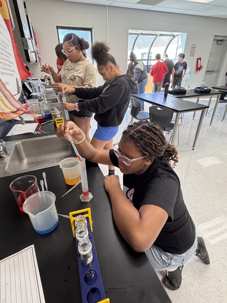 Several individuals are performing a chemistry experiment in a classroom. One is pouring liquid into a beaker.
