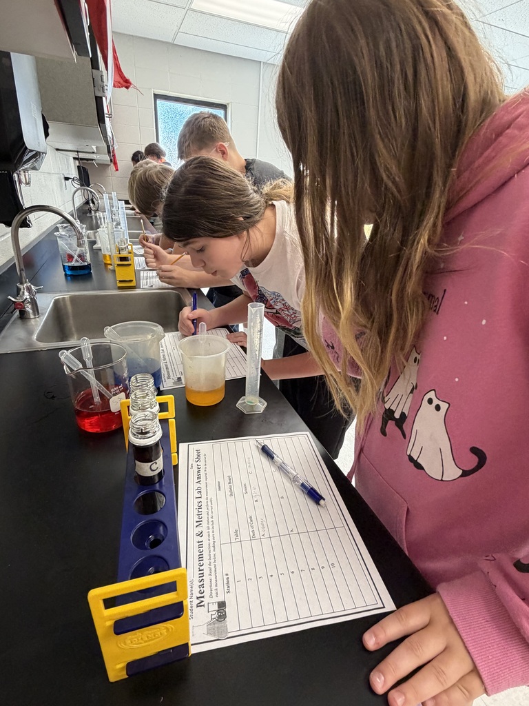 Students examine test tubes with colored liquids at a laboratory sink, with one holding a pen.