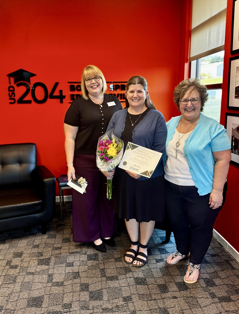 Four women pose in front of an orange wall with a graduation cap logo, holding a bouquet and certificates.