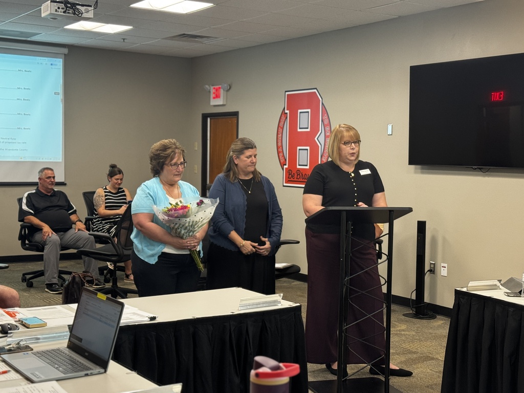 A woman speaks at a podium in a meeting room. Four people stand behind her. A screen and monitor are on the wall.
