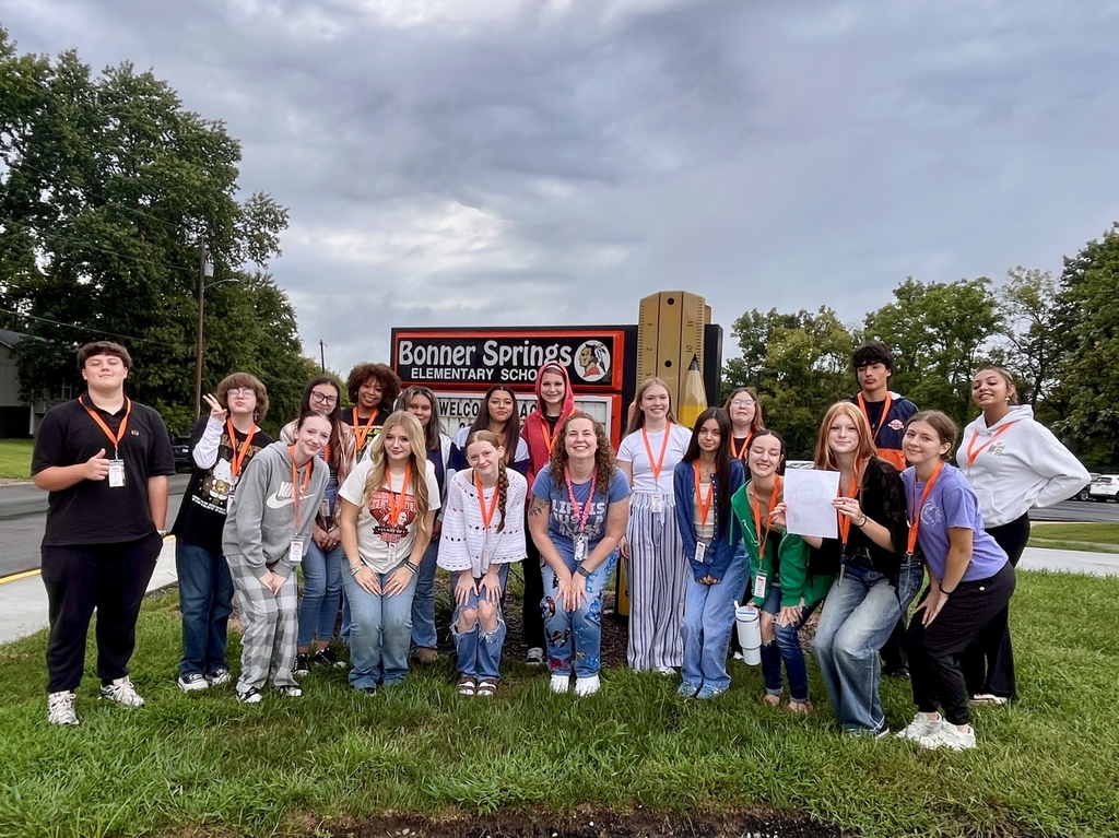 A group of students poses in front of a school sign, with some holding certificates. They stand on grass, with trees and a cloudy sky in the background.