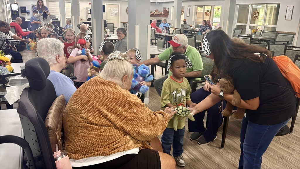 Elderly person and young child, with others, seated around tables in a dining area; woman hands child flowers.
