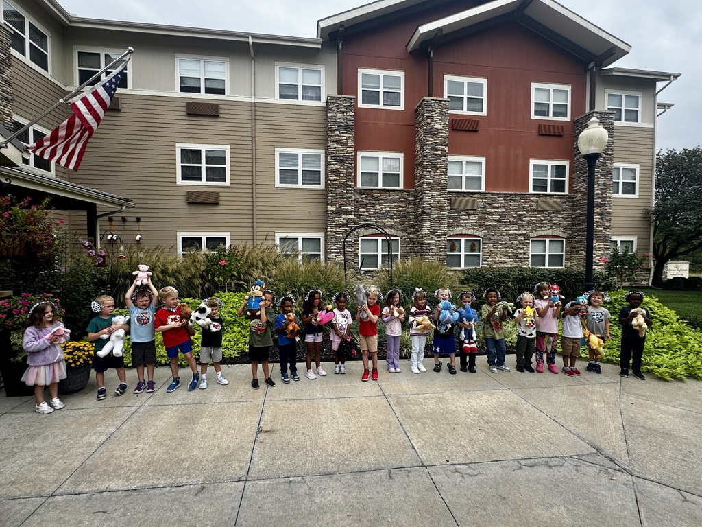 A group of children holding stuffed toys stand in front of an apartment building with an American flag.