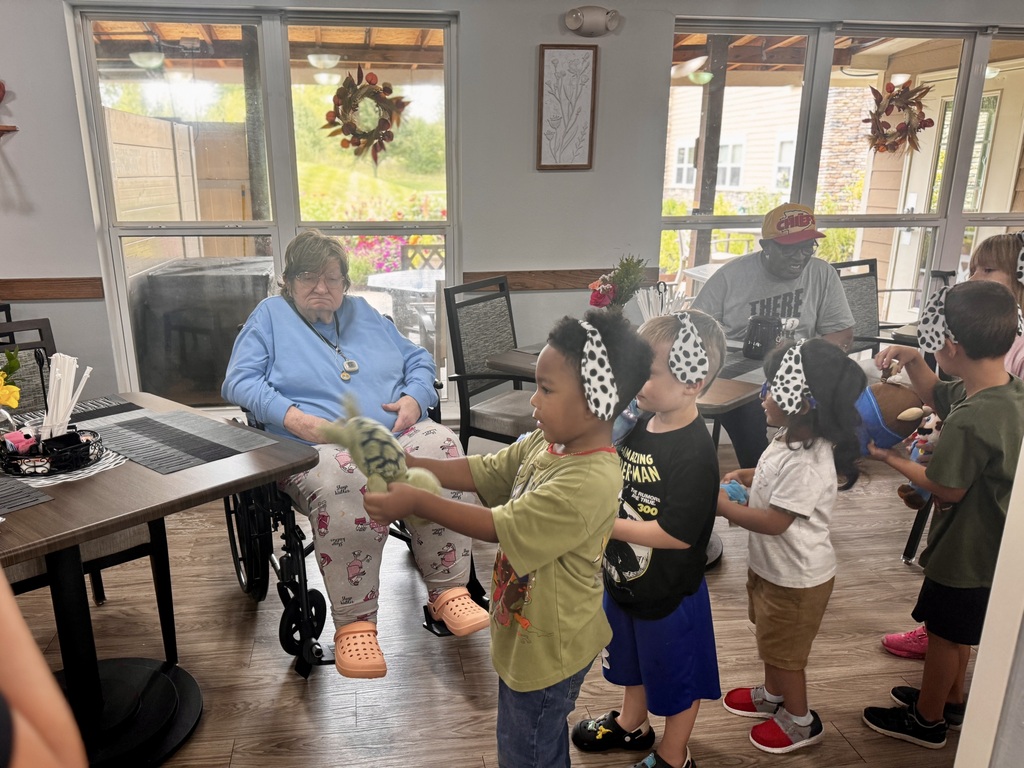 A woman in a wheelchair sits near children wearing headbands. They hold hands, while others sit at a table.