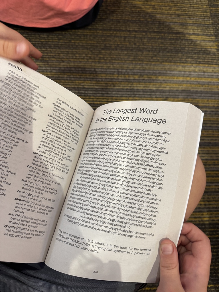 Open book displaying "The Longest Word in the English Language" in black text, held by two hands.