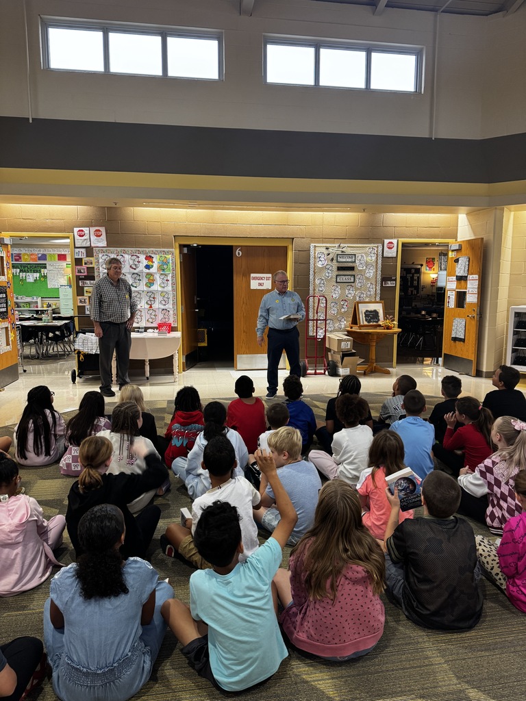 People in a room with a stage, one standing and another with a microphone, while children sit on the floor, facing the stage.