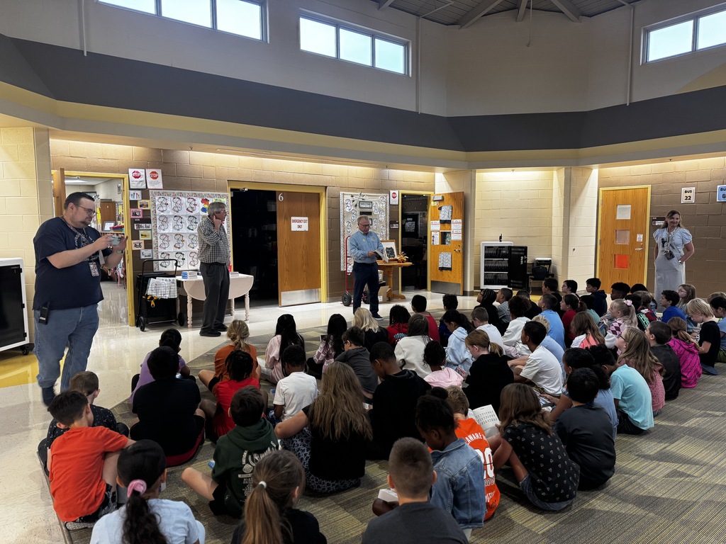 A group of children are seated in a circle in a large room, listening to two standing men.