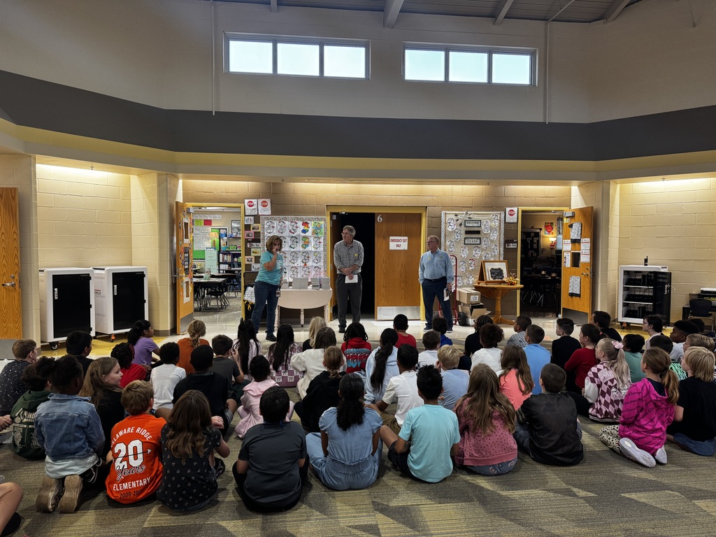 A classroom with students sitting on the floor in front of two teachers standing near a door.