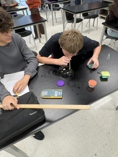 Two boys are in a classroom, one blowing into a pipette, another holding a pipette. Desks, books, and colored objects are on the table.