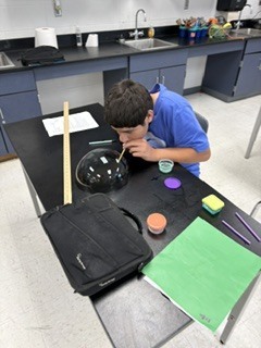 A young person in a blue shirt works at a table, dipping a stick into a jar.