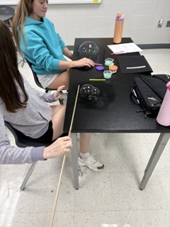 Two individuals at a table, one holding a stick and the other interacting with a glass sphere.