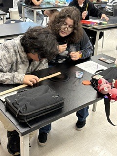 Two individuals in a classroom with a black table, one bent over a paper and the other holding a paintbrush.