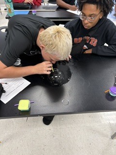 Two students in black tops working on a science project at a table; one leans over to examine a black object.