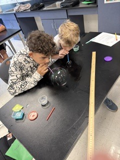 Two children are conducting a science experiment with a glass bubble, ruler, and other items.