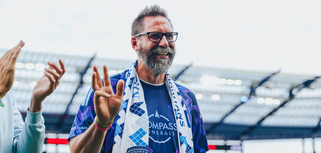 An older man in a stadium with a sign that reads "Compass Health" stands with hands raised.