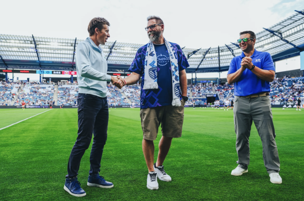Three men on a football field. One holds a handshake with another man. Stadium in the background with a crowd.